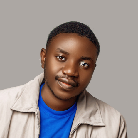 Headshot of Black man with short hair and moustache in blue t-shirt and beige coat against a grey background.