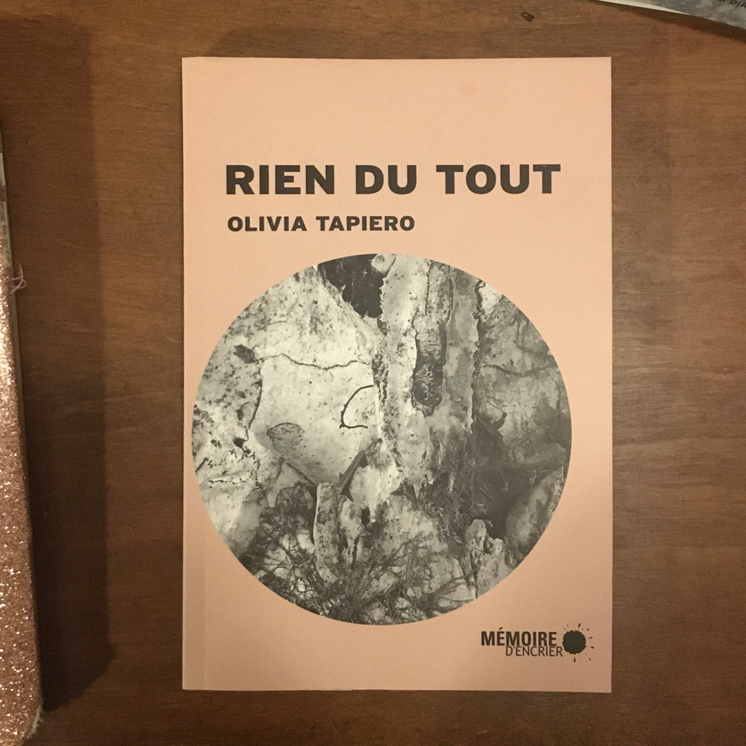 Cover of 'Rien du Tout' shot from above. Cover is peach-coloured with black font above a circle of an abstract black and white graphic. Book rests on a wooden table with other corners of books visible.