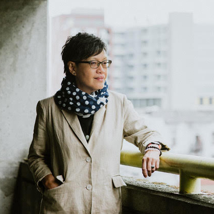Kwa in a beige linen jacket and blue polka-dotted scarf standing on a concrete balcony, arm resting on a railing, looks out on a cloudy day, with buildings obscured in the background.